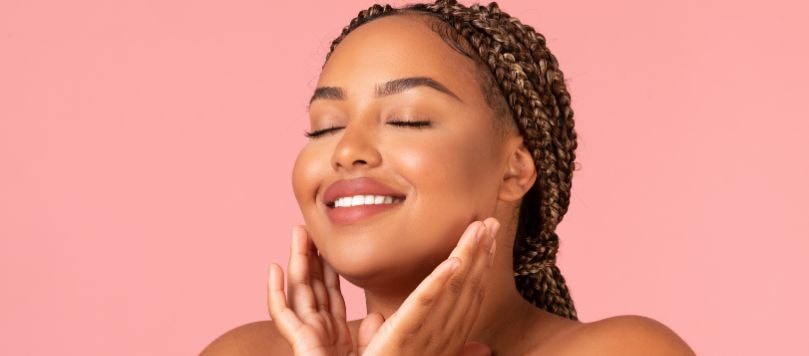 Mulher sorridente de olhos fechados, com tranças castanhas e mechas claras, tocando o rosto suavemente com as duas mãos, em frente a um fundo rosa.