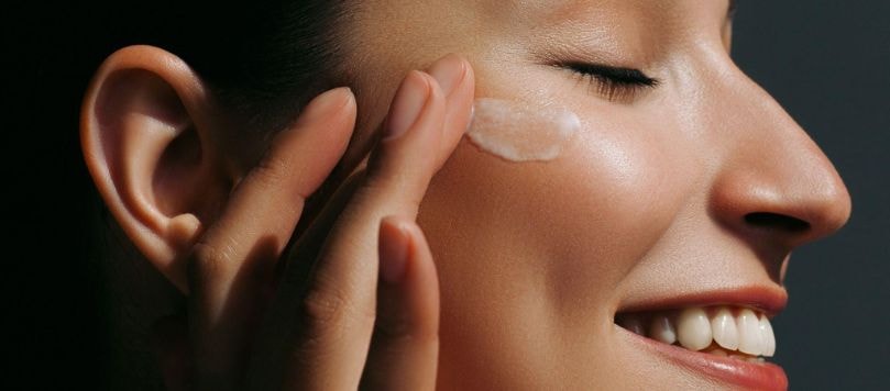Mulher sorridente aplicando creme hidratante no rosto com os dedos, com pele iluminada e saudável.