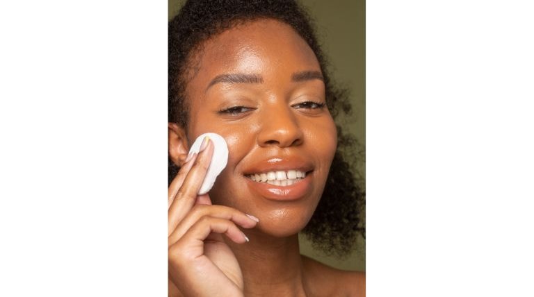 Mulher negra e sorridente aplica a água micelar com um algodão no rosto para limpar e suavizar a pele