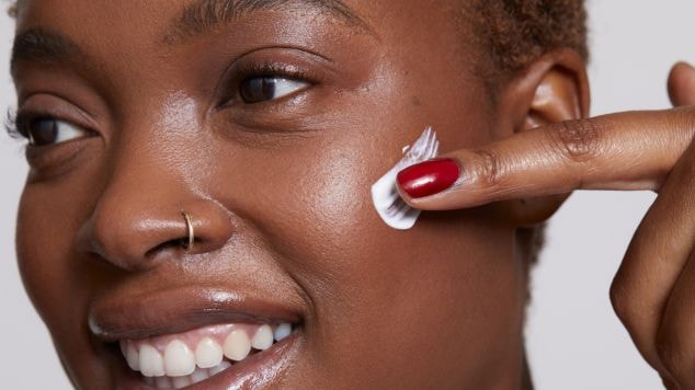 Mulher negra e sorridente aplica um creme hidratante com ácido hialurônico para o rosto.