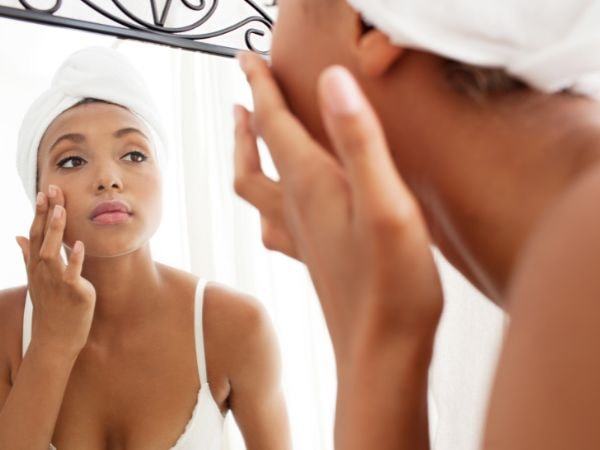 Mulher negra em frente ao espelho enquanto aplica um creme para pele do rosto em sua face.