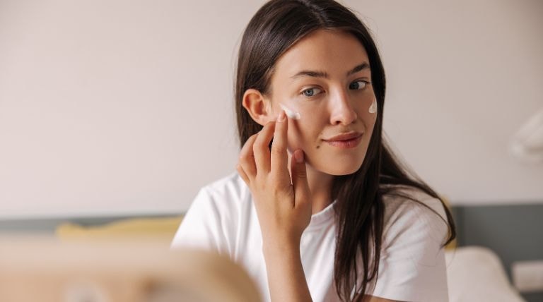 Jovem caucasiana aplica um creme para manchas no rosto em frente ao espelho