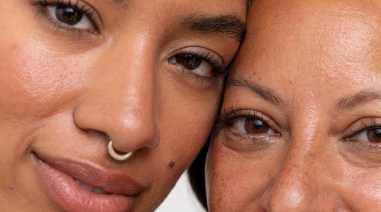 Close-up de duas mulheres com pele madura, rostos próximos em um gesto de carinho, destacando a textura natural da pele.
