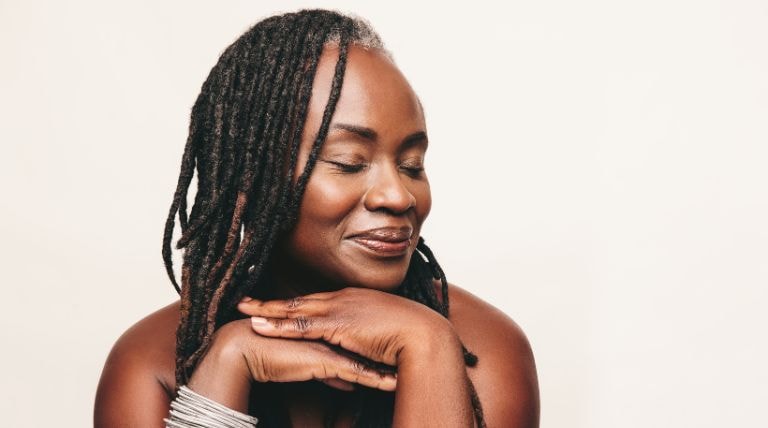Mulher negra com dreadlocks, sorrindo gentilmente com os olhos fechados e mãos sob o queixo, exibindo sua pele madura e radiante.