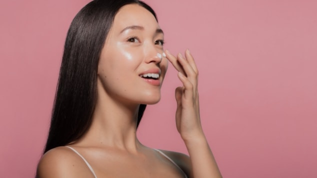 Mulher com pele luminosa aplicando pontos de hidratante no rosto com os dedos, demonstrando a técnica de skincare em camadas.
