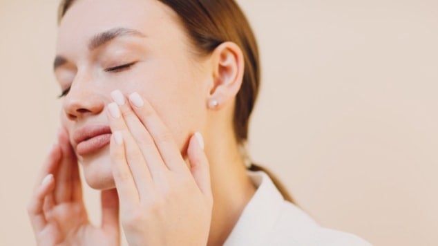 Mulher de olhos fechados toca suavemente o rosto com expressão serena, exibindo pele luminosa e bem cuidada em fundo neutro.