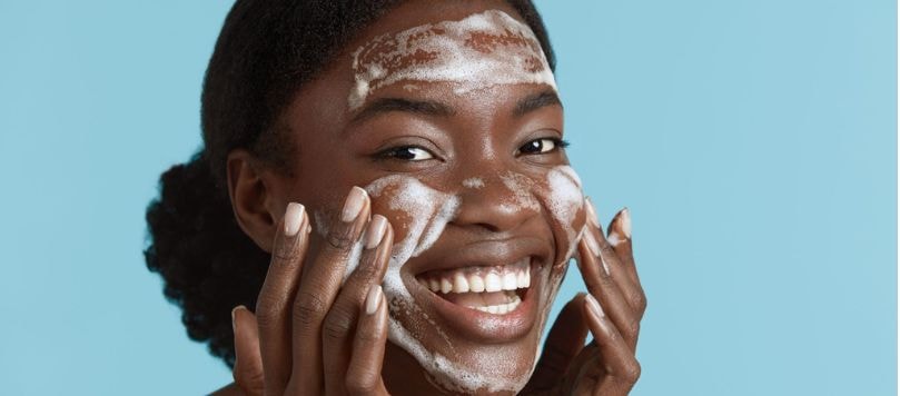 Mulher sorridente com espuma de sabonete no rosto, realizando a limpeza facial com as mãos, transmitindo leveza e bem-estar na rotina de skincare.