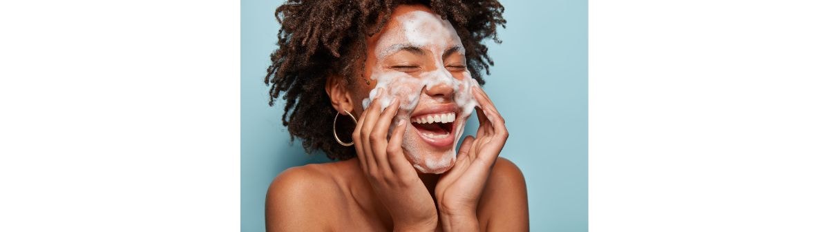  Close-up de uma mulher com cabelos escuros cacheados, sorrindo com os olhos fechados enquanto massageia o rosto com espuma de limpeza e as mãos, em um fundo azul.