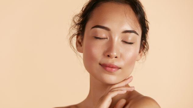 Mulher de olhos fechados, cabelo castanho preso, apoiando o rosto nas mãos em um fundo rosa.