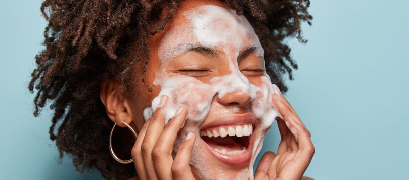  Mulher jovem de cabelos cacheados ri enquanto lava o rosto com espuma de limpeza, demonstrando prazer e leveza na rotina de skincare.