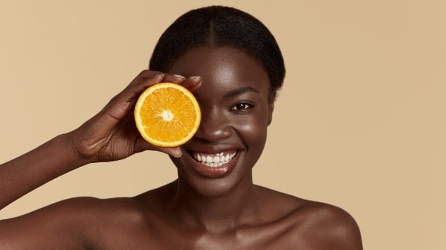 Mulher sorrindo com o cabelo preso e a metade de uma laranja tampando o seu olho direito em um fundo bege.